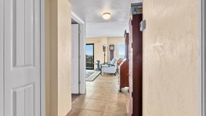 A hallway with a white door, beige walls, and a tiled floor leading to a living area with a white sofa and a wooden cabinet. - Direct Beach Views - Sleeps 4, Corner Unit Tennis - Pool (Flagler Beach)