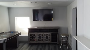 A compact living space with a flat-screen TV mounted on the wall, a small bar area with stools, and a window providing natural light. - Spacious Cheerful 1-Bedroom Residential Home (Tulsa)