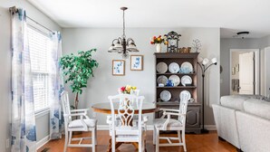 Un coin repas avec une table ronde, des chaises blanches, un lustre, une plante en pot et un porte-assiettes fixé au mur.