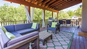 Een terras buiten met een houten dek, een blauwe bank met groene kussens, een houten tafel en een barbecue.