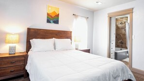 A neatly made bed with white bedding, two bedside lamps, a wooden nightstand, a wall-mounted painting, and a bathroom with a shower and a tiled wall. - Spacious Retreat w/ Views Near Zion & Bryce UT (Kanab)