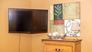 A television mounted on a wall, a wooden cabinet with a lamp, and a decorative wall piece. - Vacation Home w/red rock views, Near Zion & Bryce (Kanab)