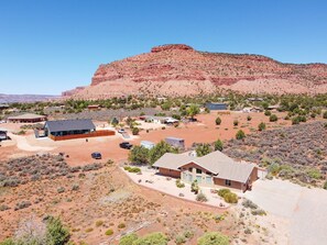 Paisaje desértico con una gran formación rocosa roja al fondo, rodeado de vegetación escasa y varias edificaciones con techos planos y diseños sencillos.