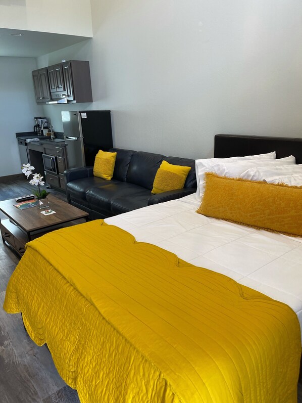 Moderni hotellihuone, jossa on suuri sänky, musta nahkasohva, pieni keittokomero ja puinen sohvapöytä.
