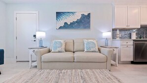 A living room with a beige sofa, two matching pillows, a rug, a painting, a kitchen area with cabinets and a stove, and a door.