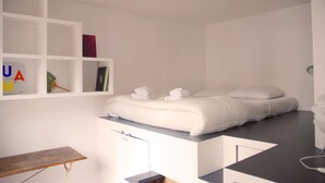 A minimalist hotel room with a bed, a small table, and a shelf with books and decorative items. - ★ Zen cocoon, calm & bright in Belleville ★ (Paris)