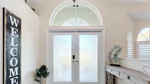Un vestíbulo amplio con puerta de vidrio satinado, un letrero de bienvenida, una planta en maceta y una consola con objetos decorativos.