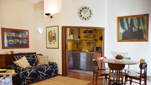 Una sala de estar con un sofá, una mesa de comedor con sillas, una cocina al fondo y un reloj en la pared.