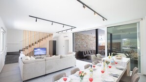 A modern living room with a sofa, dining table set for a meal, and a TV on a stand. - Ayia Napa Kube Villa KB4 -  a villa that sleeps 12 guests  in 5 bedrooms (Ayia Napa)