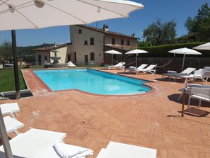 Una piscina con sillas de descanso y sombrillas, rodeada por un patio de ladrillo y un edificio al fondo.