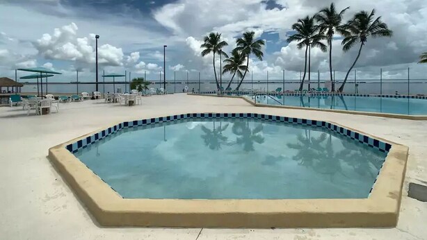 Ein Pool mit einer gefliesten Kante, umgeben von Liegestühlen und Sonnenschirmen, unter einem bewölkten Himmel.
