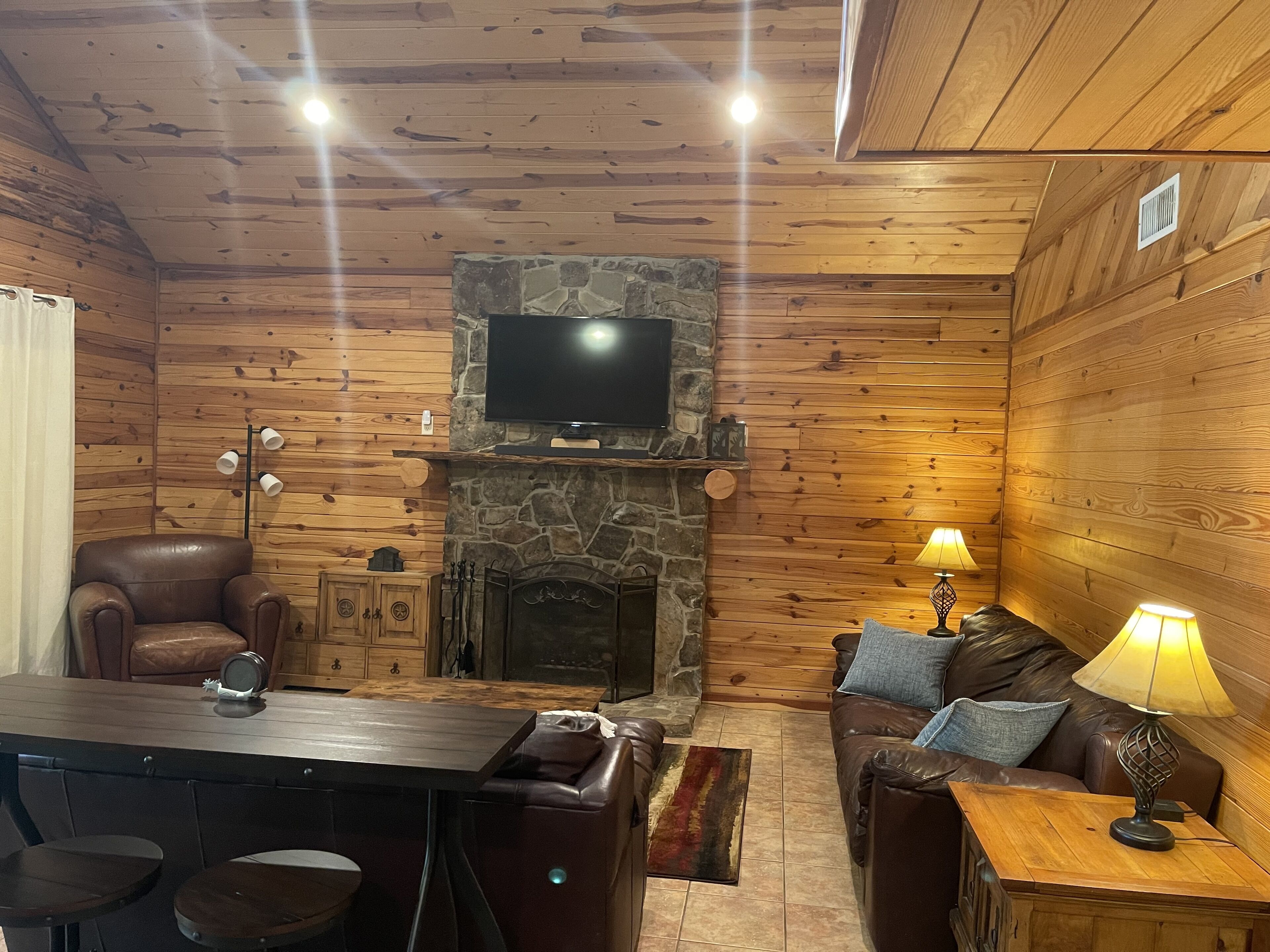 A cozy cabin interior with a stone fireplace, wooden walls, a leather sofa, a dining table with chairs, and a television mounted above the fireplace.
