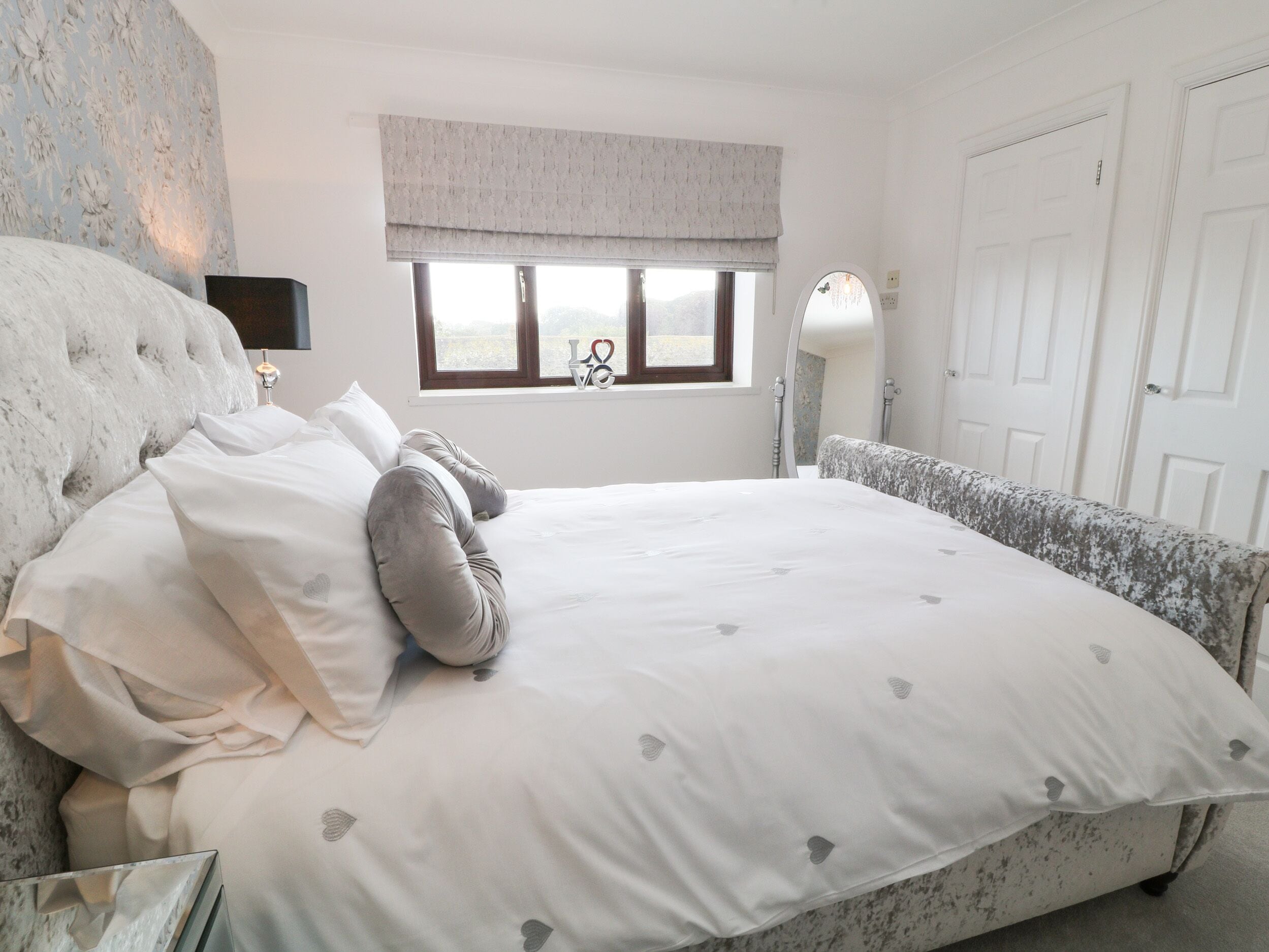 A bedroom with a large bed, a window with a view, a bedside lamp, and a door.