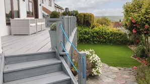 Una escalera de madera que conduce a una terraza con mobiliario exterior, un césped cuidado y vistas al mar.