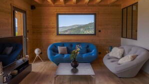 A cozy living room with a wooden ceiling, a large window with a view of a green landscape, a blue sofa, a grey armchair, and a small table with a vase. - Chalet Les P'tits Biquets  (GERARDMER)