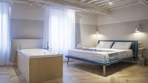 A modern bedroom with a wooden floor, a large bathtub, a bed with blue headboard, and two bedside lamps.