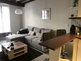 A modern living room with a grey sofa, a wooden coffee table, and a kitchen area with a bar counter and coffee machine.