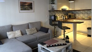 A modern living room with a grey sofa, a coffee table with remote controls, and a kitchen area with a microwave and a sink. - Charmant Zola (Troyes)