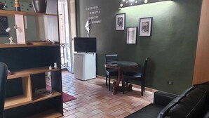 Un espacio habitable compacto con sofá, una pequeña mesa de comedor y un refrigerador. Hay cuadros enmarcados en la pared y un estante con varios objetos.