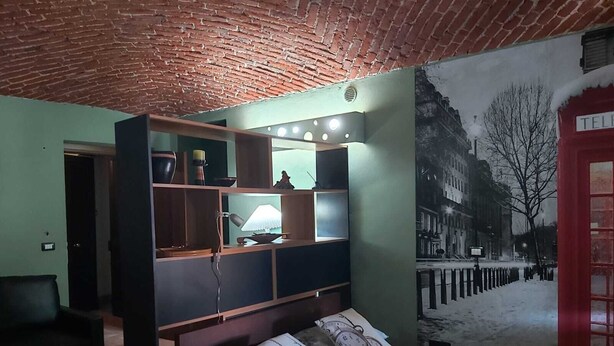 Un dormitorio con techo de ladrillo, cama con almohadas estampadas, mesita de noche y un mural en la pared que muestra una calle nevada.