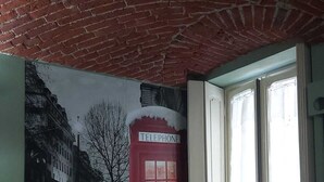 Una habitación con techo de ladrillo, un ventanal con cortinas, una cama con cabecera blanca y un mural de un teléfono rojo.
