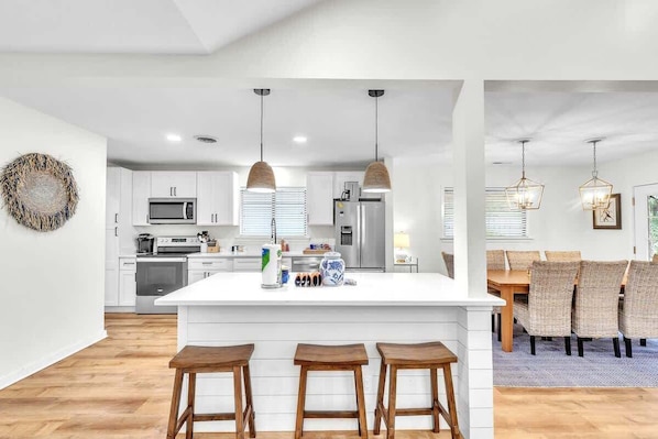 A modern kitchen with white cabinetry, a central island with stools, pendant lighting, and a dining area with wicker chairs.