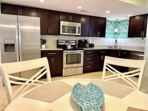 A modern kitchen with dark wood cabinets, stainless steel appliances, and a white dining table with a blue woven centerpiece.