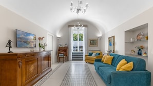 A spacious living room with a wooden cabinet, a blue sofa with yellow cushions, a chandelier, and a large window with a view of greenery. - SMALL COURT MAISON (Piano di Sorrento)