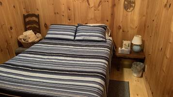 A wooden cabin room with a bed, a nightstand, a basket of towels, a lamp, and a wall decoration.