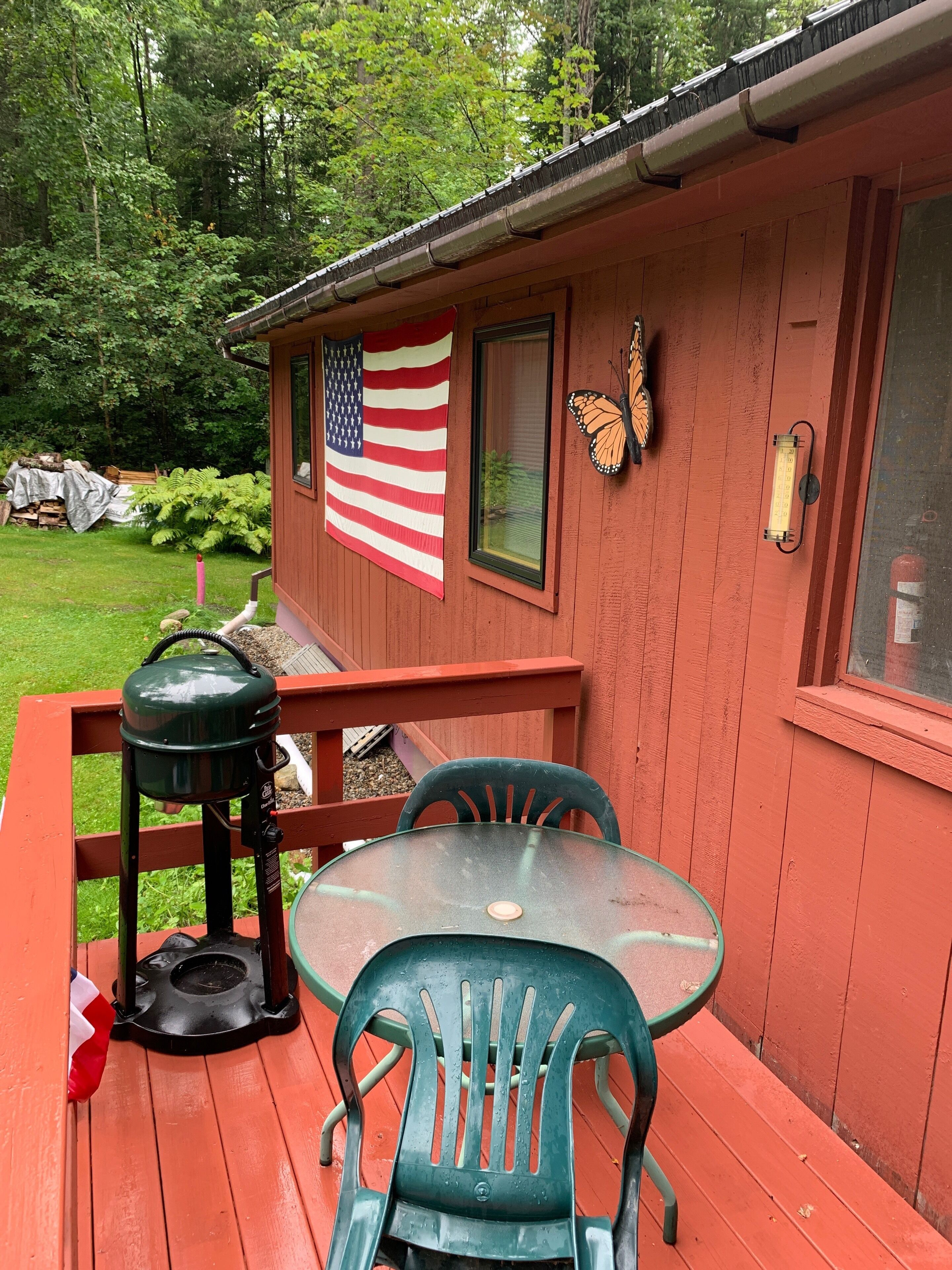 En træhytte med rødt ydre, et glasbord, to grønne stole, en grill og en sommerfugludsmykning, samt et amerikansk flag.