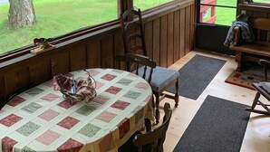 A wooden cabin with a table covered by a quilt, a rocking chair, and a view of a green landscape through large windows. - Cozy cottage (Lunenburg)