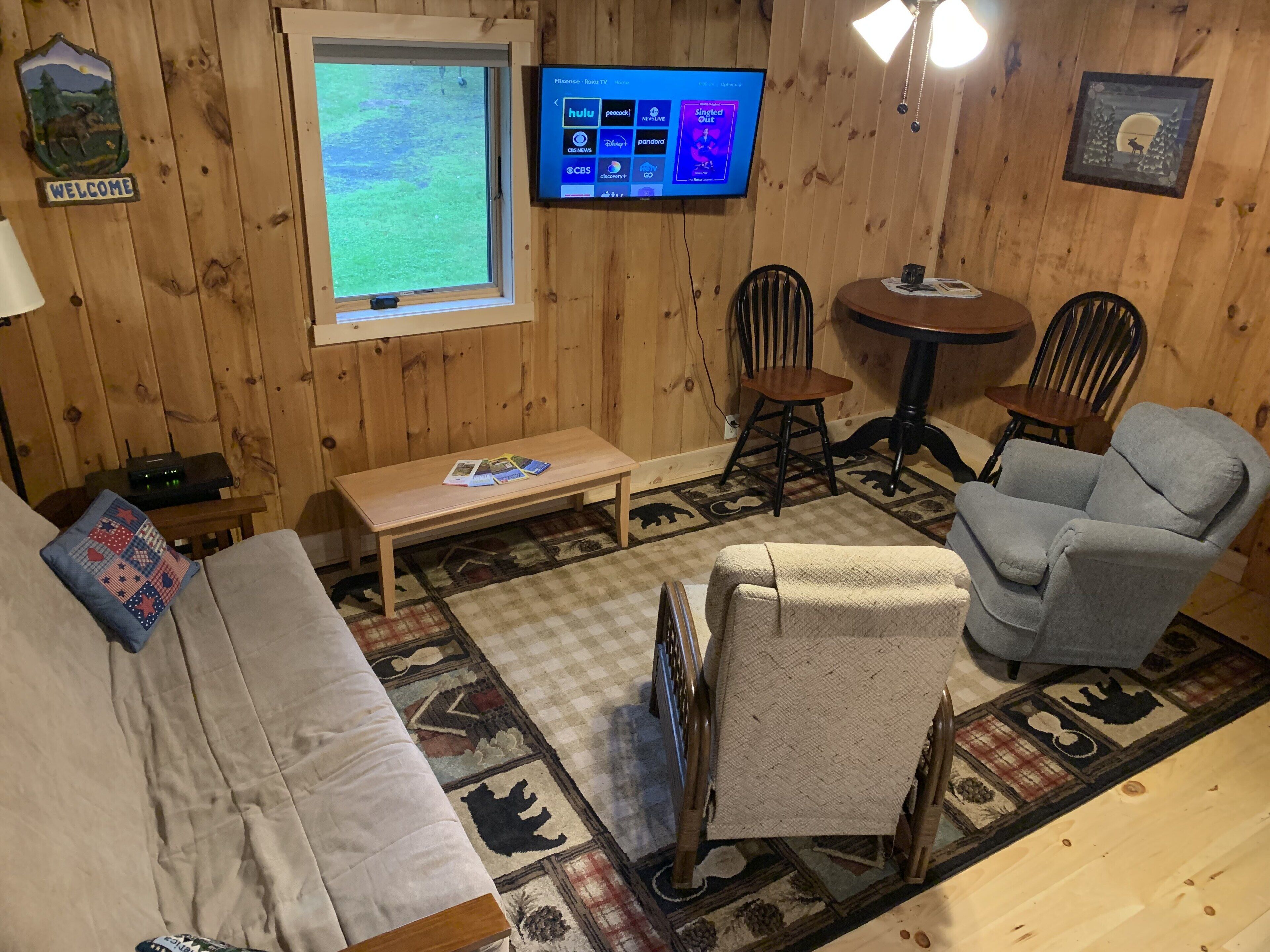 Een gezellig chaletinterieur met houten inrichting, een aan de muur bevestigde televisie, een bank, een stoel en een kleine tafel.