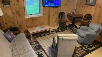 Een gezellig chaletinterieur met houten inrichting, een aan de muur bevestigde televisie, een bank, een stoel en een kleine tafel.