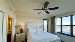 Una habitación de hotel con cama, mesita de noche, lámpara, ventilador de techo y un ventanal con vista al mar.