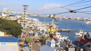 Una persona haciendo tirolesa sobre un puerto deportivo con barcos y edificios al fondo.