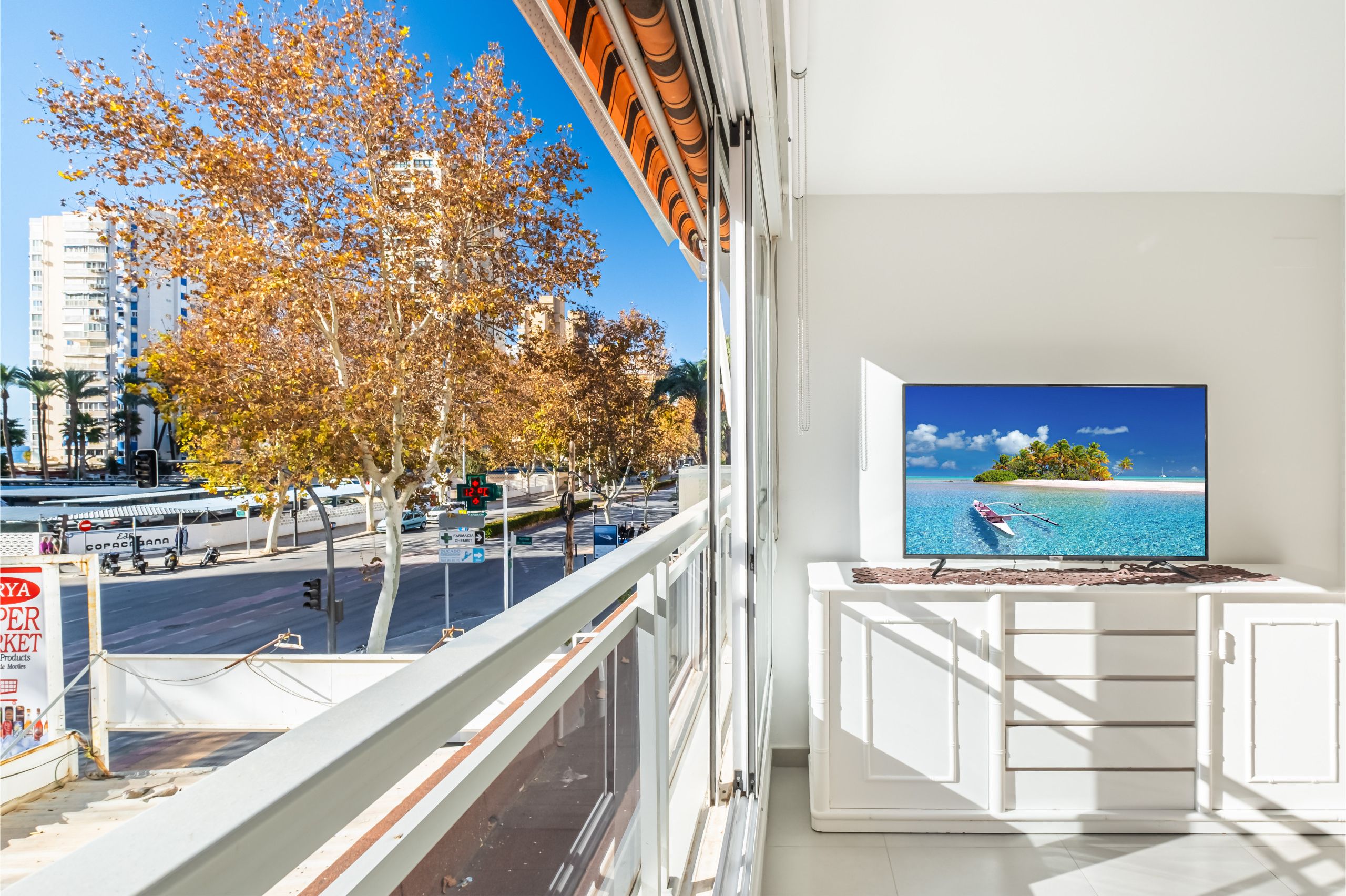 A balcony with a TV displaying a beach scene, overlooking a street with autumn trees and buildings.