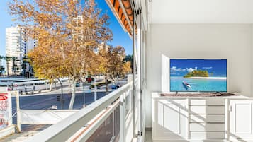 A balcony with a TV displaying a beach scene, overlooking a street with autumn trees and buildings.