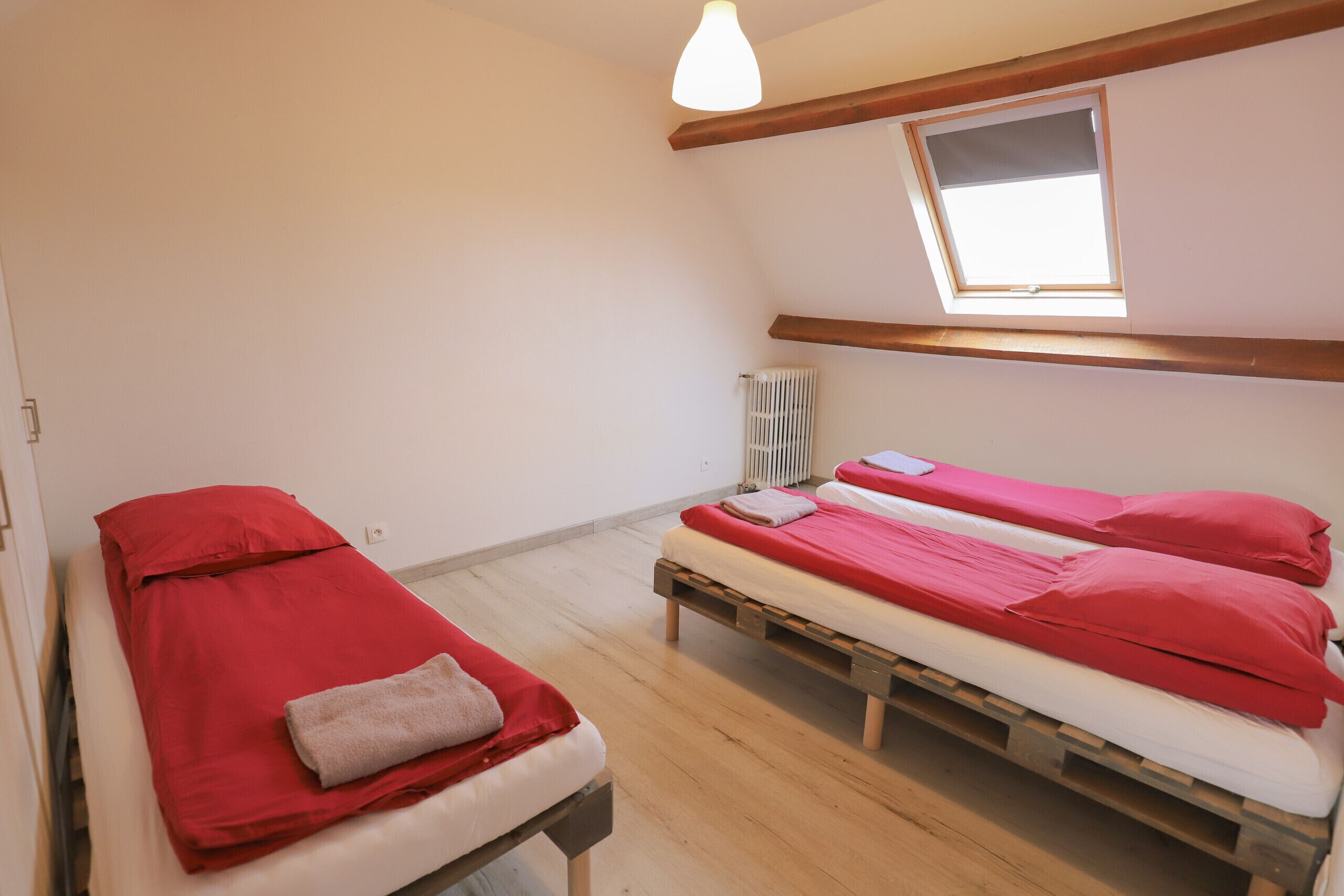 Habitación con dos camas, cada una con ropa de cama roja y cabeceros de madera. Hay un tragaluz sobre las camas, un radiador en la pared y una puerta blanca a la izquierda.