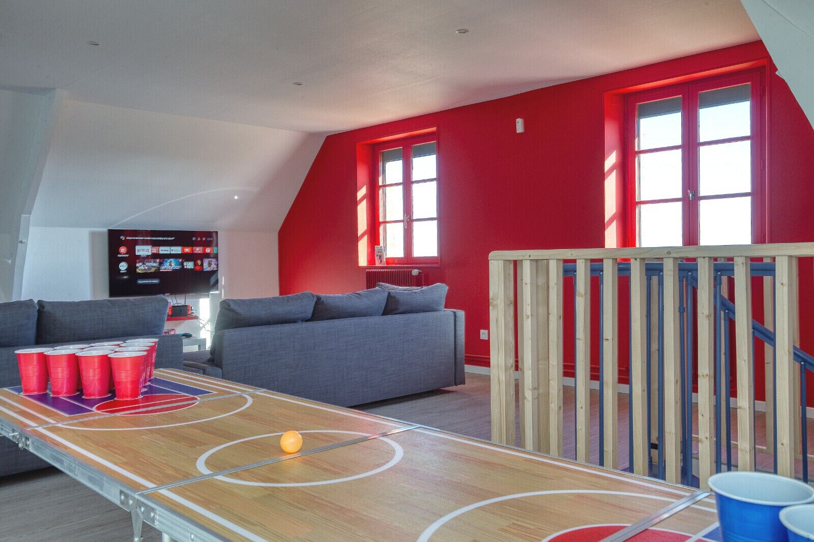 Un salon avec un mur rouge, un canapé gris, une table basse en bois et un téléviseur fixé au mur.