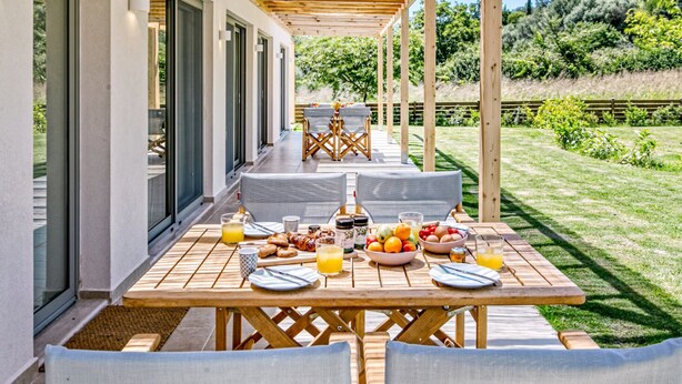 Un espace extérieur abrité, aménagé pour les repas, avec une pergola en bois, une table dressée avec des assiettes, des verres et des fruits, et une vue sur un jardin luxuriant.