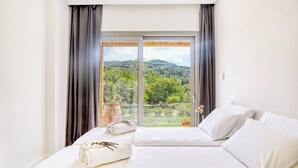 Une chambre d’hôtel avec un lit, du linge de lit blanc, une vue sur un paysage verdoyant par une grande fenêtre, et un vase rempli de fleurs posé sur le rebord de la fenêtre.