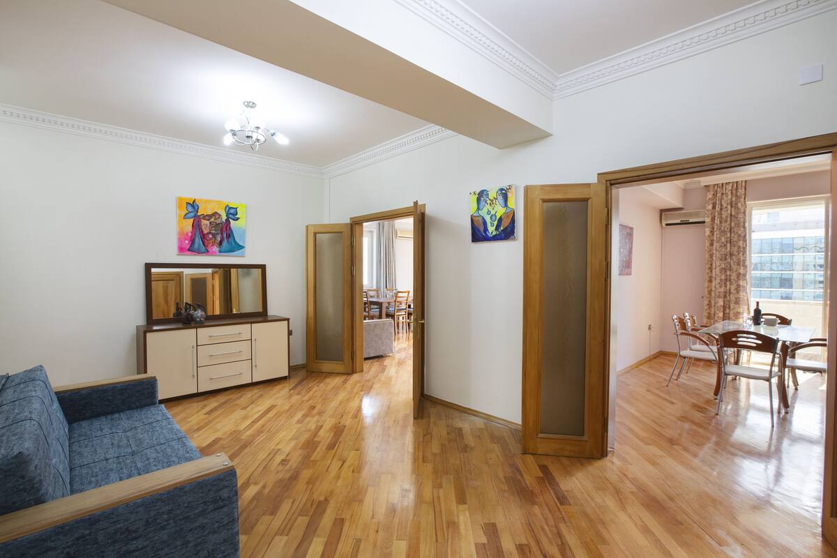Un salon spacieux doté d’un plancher en bois, d’un canapé bleu, d’un coin repas avec une table et des chaises, et de deux tableaux accrochés aux murs.