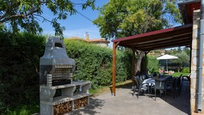 Een terras buiten met een stenen barbecue, een overdekte zithoek en uitzicht op een woonwijk.