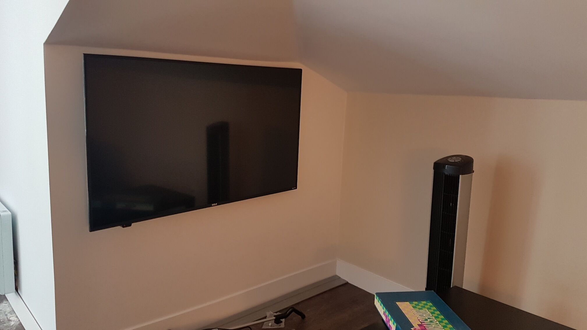 A room with a large flat-screen TV mounted on the wall, a black standing fan, and a small table with a book.