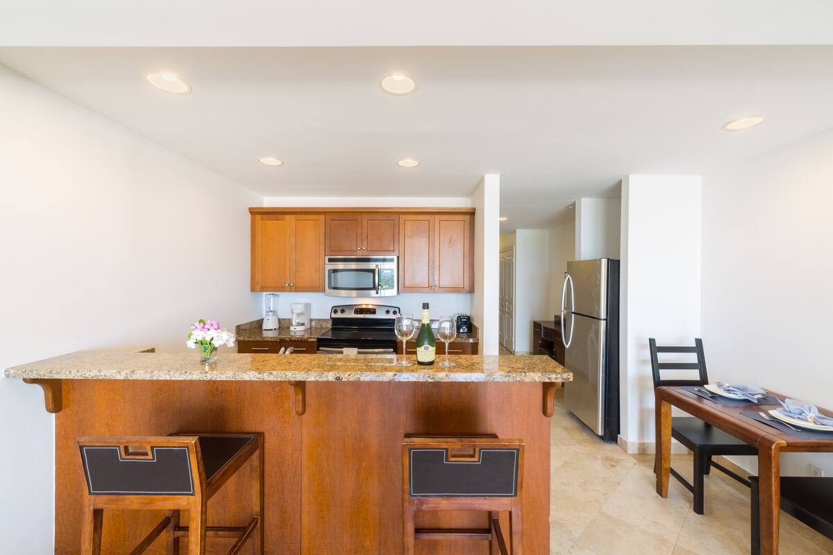 A modern kitchen with wooden cabinets, a granite countertop, stainless steel appliances, and a bar area with stools.