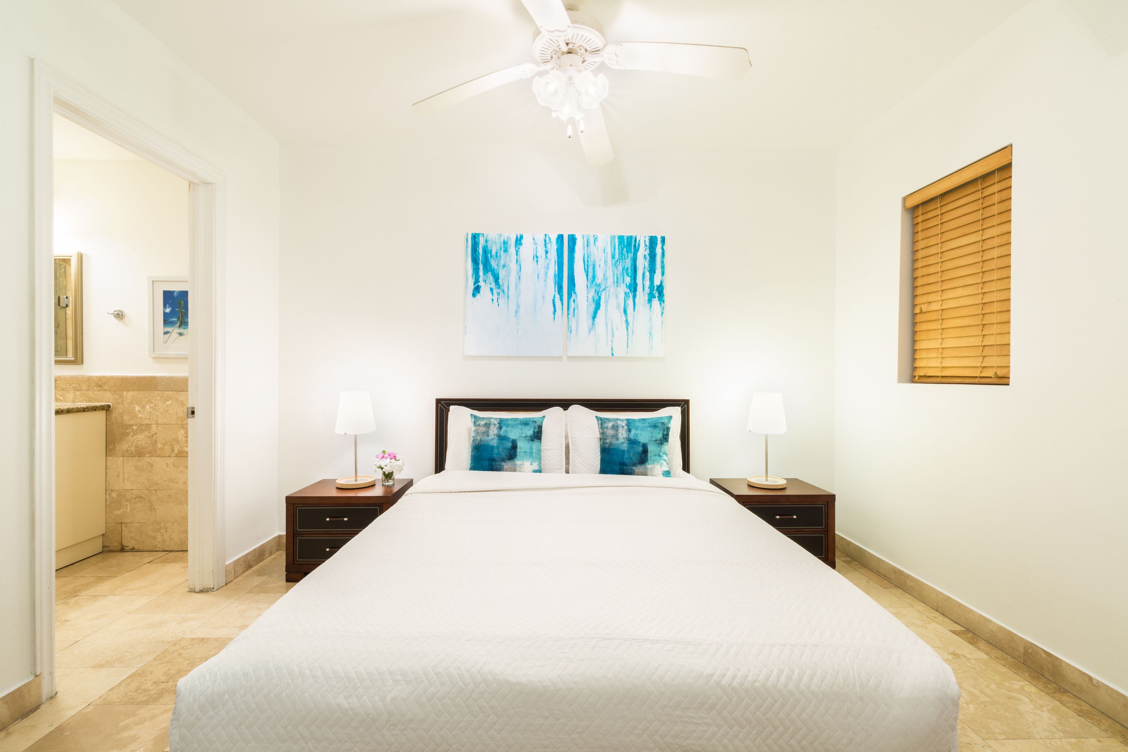 A neatly arranged bedroom with a large bed, two bedside tables with lamps, a ceiling fan, a wall-mounted artwork, and a window with blinds.