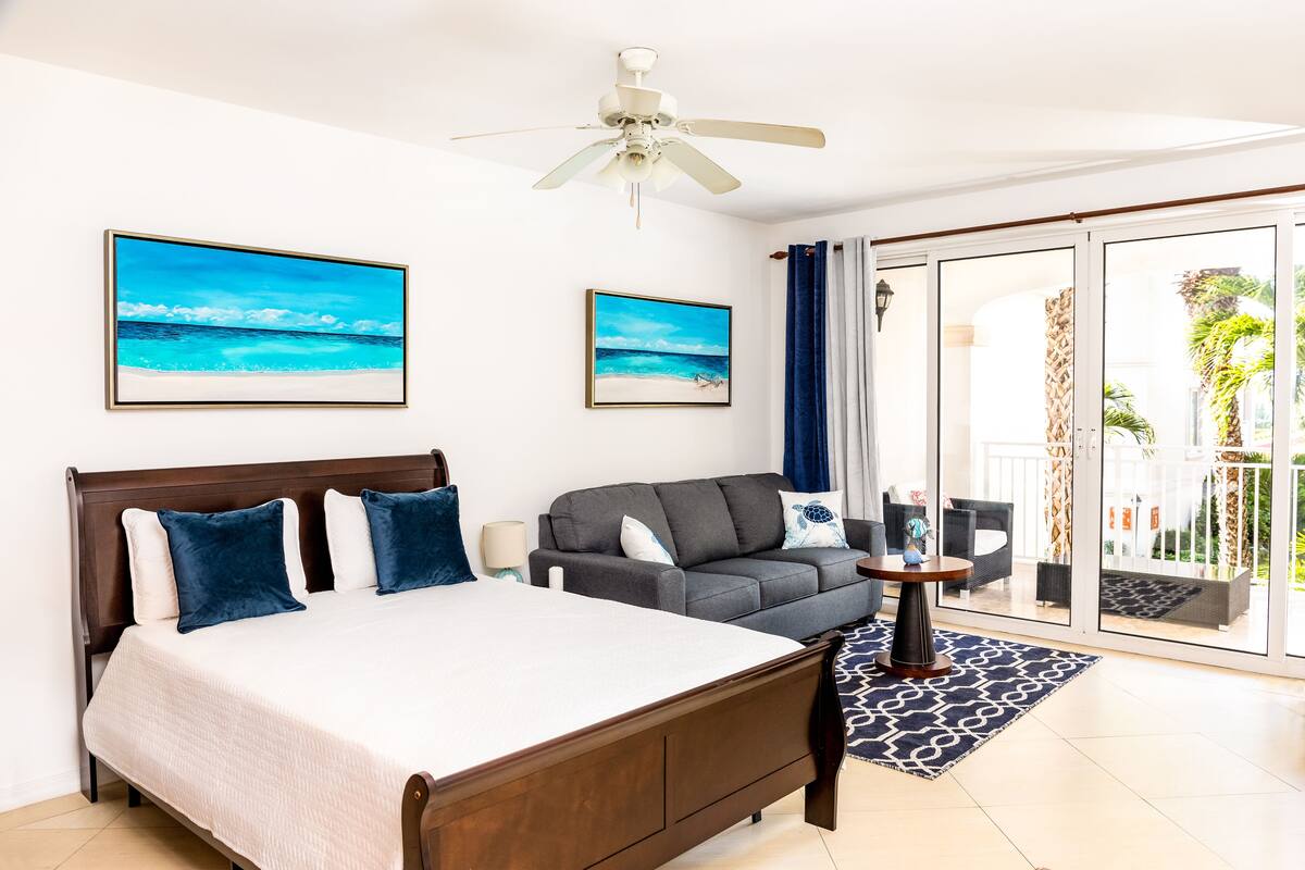 A bedroom with a bed, a sofa, a ceiling fan, and two framed pictures of a beach.
