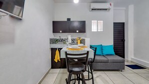 Une cuisine compacte avec un coin repas, un canapé gris agrémenté d’un coussin bleu et un téléviseur fixé au mur.