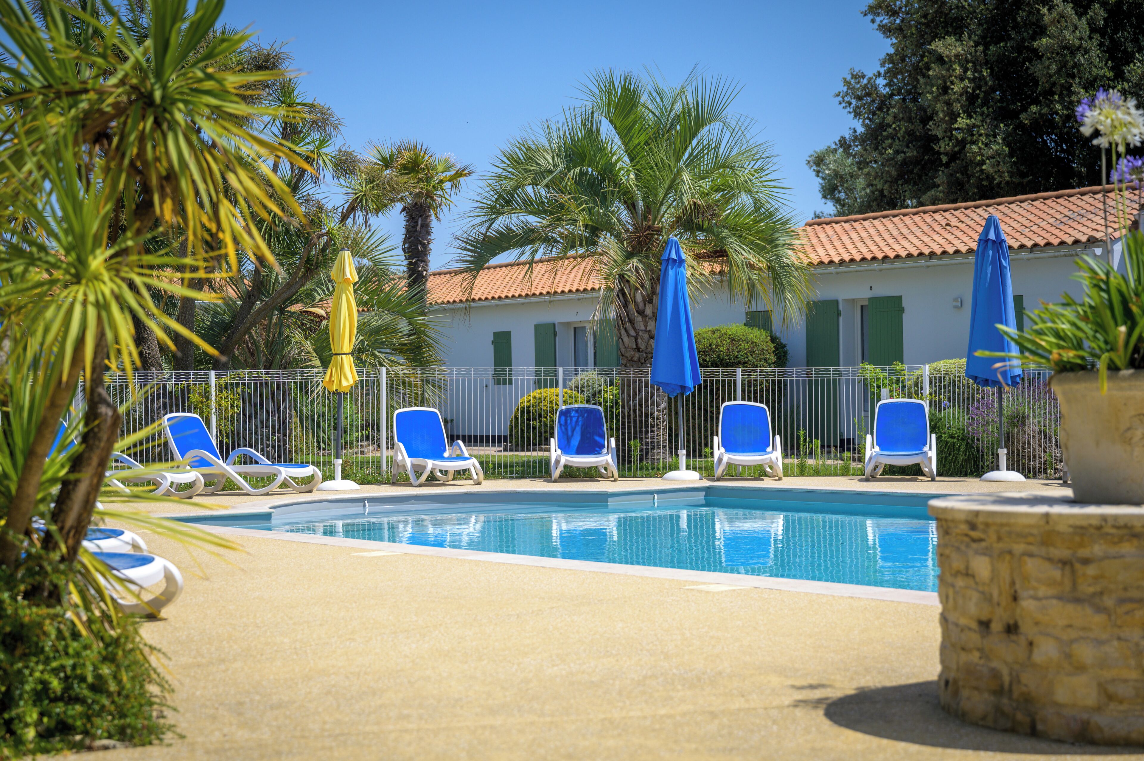 A pool area with blue lounge chairs and umbrellas, surrounded by palm trees and a building with a tiled roof.