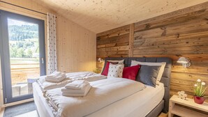 A bedroom with a large bed, wooden panel walls, a view of the outdoors, and a small bedside table with a lamp and a flower vase.